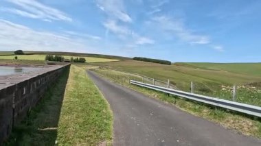 İskoç sınırlarındaki su deposu barajının yanında yürürken beton duvar boyunca uzanan yollar hala su ve açık yaz gökyüzünün altındaki bozkır tepelerini yansıtıyor..
