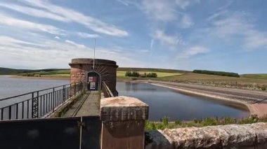 İskoç sınırlarındaki su deposu barajının yanında yürürken beton duvar boyunca uzanan yollar hala su ve açık yaz gökyüzünün altındaki bozkır tepelerini yansıtıyor..