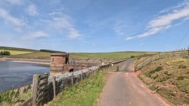 İskoç sınırlarındaki su deposu barajının yanında yürürken beton duvar boyunca uzanan yollar hala su ve açık yaz gökyüzünün altındaki bozkır tepelerini yansıtıyor..