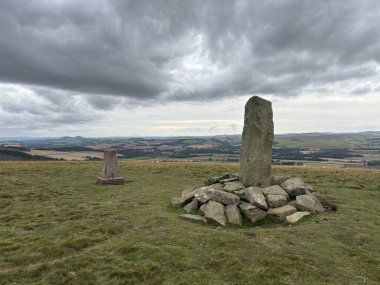Upright ancient stone marking the center of Iron Age hillfort on Dabshead Hill in Scottish Borders, overlooking open moorland with heather, grassy slopes, sheep pastures, winding streams, and layered hills under clear sky.
