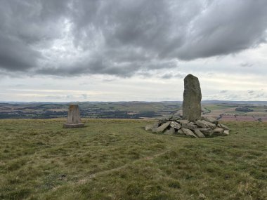 Upright ancient stone marking the center of Iron Age hillfort on Dabshead Hill in Scottish Borders, overlooking open moorland with heather, grassy slopes, sheep pastures, winding streams, and layered hills under clear sky.
