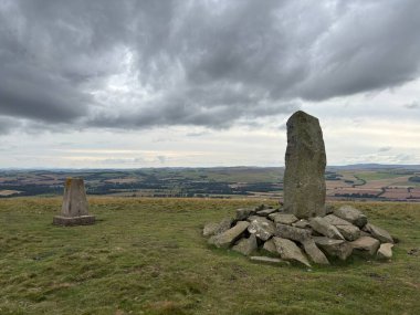 Upright ancient stone marking the center of Iron Age hillfort on Dabshead Hill in Scottish Borders, overlooking open moorland with heather, grassy slopes, sheep pastures, winding streams, and layered hills under clear sky.