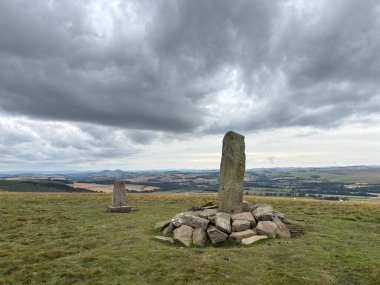 Upright ancient stone marking the center of Iron Age hillfort on Dabshead Hill in Scottish Borders, overlooking open moorland with heather, grassy slopes, sheep pastures, winding streams, and layered hills under clear sky.