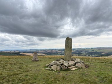 Upright ancient stone marking the center of Iron Age hillfort on Dabshead Hill in Scottish Borders, overlooking open moorland with heather, grassy slopes, sheep pastures, winding streams, and layered hills under clear sky.