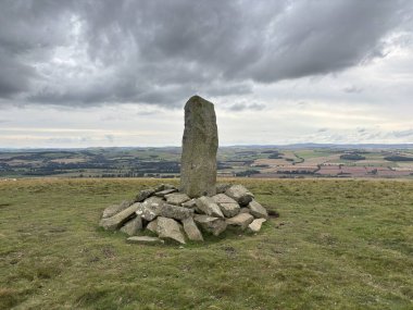 Upright ancient stone marking the center of Iron Age hillfort on Dabshead Hill in Scottish Borders, overlooking open moorland with heather, grassy slopes, sheep pastures, winding streams, and layered hills under clear sky.