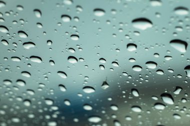 Üzerinde yağmur damlaları olan bir pencere. Yağmur damlaları küçük ve dağınık, bulanık bir etki yaratıyor. Manzara sakin ve huzurlu, yağmur damlaları pencereye yavaşça düşerken