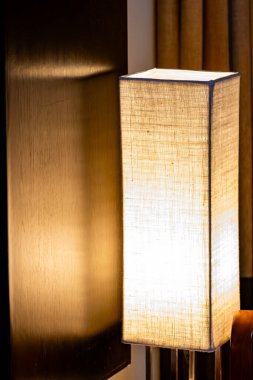 Beyaz gölgeli bir lamba parlıyor, duvara gölge düşürüyor. Işık sıcak ve davetkar. Odada rahat bir atmosfer yaratıyor.