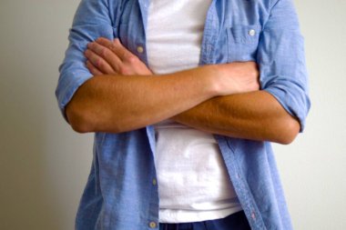 Kollarını kavuşturmuş beyaz tişörtlü ve katlanmış mavi gömlekli bir adam..