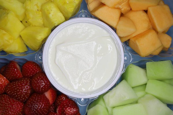 a platter with fresh fruit pieces: pineapple, strawberries, green and orange melon, sauce in the center, arranged in a disposable container with five sections