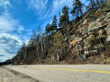 Ağaçlar ve açık mavi gökyüzü ile çevrili kayalık bir tepenin dibinde asfalt bir yol..