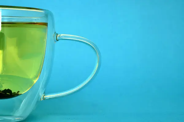 Parlak mavi arka planda yeşil çayla dolu bir cam bardak. Şeffaf tasarım içkinin ışığını ve gölgesini vurgular..