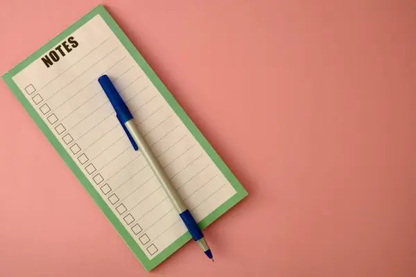 Yeşil çerçeveli bir not defteri ve yanında mavi bir kalem bulunan pembe arka planda duran sözcük notları. Planlama için minimalist kompozisyon.