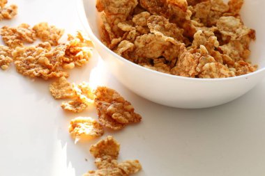 Beyaz bir masanın üzerinde gevrek mısır gevreği kasesi ve etrafa yayılmış gevrekler. Güneş ışığı yumuşak gölgeler yaratır.