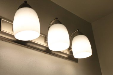 Üç duvara monte edilmiş cam gölgeli ışıklar bej bir duvara sıcak ışık yayar..