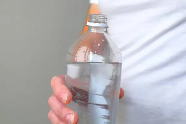Beyaz tişörtlü bir insan, yüzeyinde yoğunlaşma damlacıkları olan açık plastik bir su şişesi tutar.