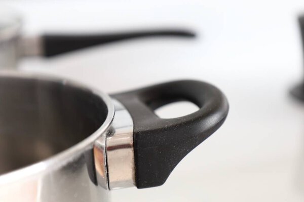 a close-up of a black plastic handle on a stainless steel pot. the handle is attached with a metal bracket. the background is blurred, showing another pot handle.