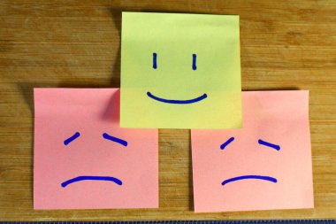 Üzgün ve kızgın pembe yüzler arasında çizilmiş yapışkan notlar. Olumsuzluklar arasında pozitifliği sembolize eden bir sarı var.