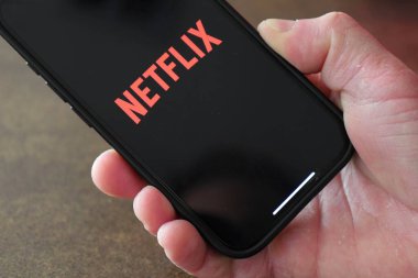 Netflix uygulaması açık olan akıllı telefonu tutan bir el, kırmızı logolu siyah ekran, bulanık arkaplan.