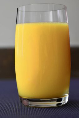 Mavi bir masa örtüsüne yerleştirilmiş, içi taze sarı-portakal suyuyla dolu şeffaf bir bardağın yakın çekimi..