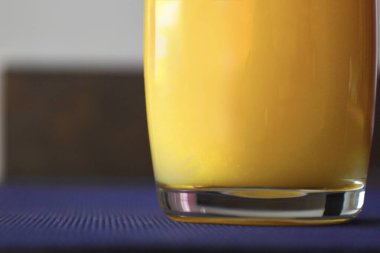 Mavi bir masa örtüsüne yerleştirilmiş, içi taze sarı-portakal suyuyla dolu şeffaf bir bardağın yakın çekimi..