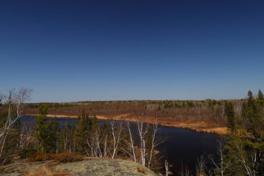 Sakin bir nehir, kuzeydeki açık mavi gökyüzünün altında kayaların ve çam ağaçlarının arasından akar..