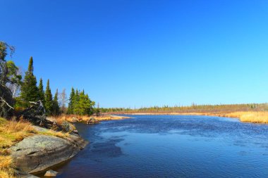 Sakin bir nehir, kuzeydeki açık mavi gökyüzünün altında kayaların ve çam ağaçlarının arasından akar..