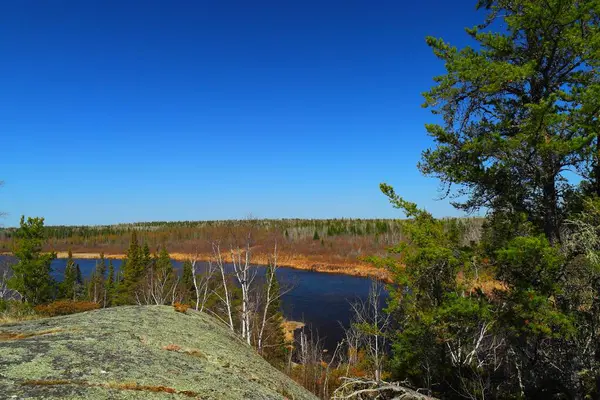 Sakin bir nehir, kuzeydeki açık mavi gökyüzünün altında kayaların ve çam ağaçlarının arasından akar..