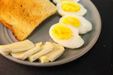 Kızarmış ekmek, kaynamış yumurta kremalı yumurta ve gri tabakta bir dilim yumuşak peynir..