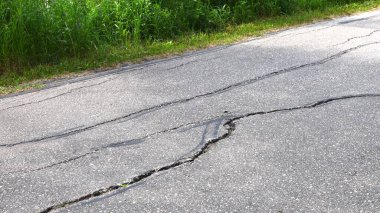 Hasarlı asfalt yol yüzeyinde görünür çatlaklar ve yazın yol kenarında yeşil çimenler.