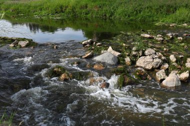 Yeşil çayırlar boyunca akan küçük bir nehir ya da akarsu. Köpüklü su yosunlarla kaplı taşlar arasında akar, sakin ve yansıtıcı yüzeyle dinamik bir kontrastlık yaratır.. 