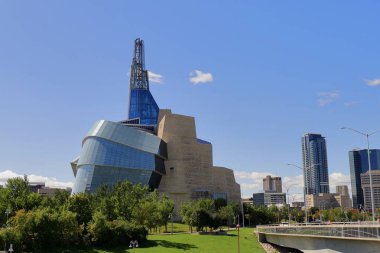 Kanada İnsan Hakları Müzesi Winnipeg, Manitoba, Kanada. Cam ve taş tasarımlı modern mimari, yeşil park ve mavi gökyüzünün altındaki şehir silueti.