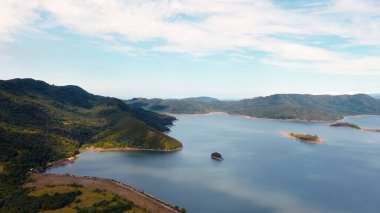 Yüksek dağlarla çevrili yemyeşil bir vadide yuva yapmış dingin bir gölün nefes kesici 4K drone görüntüsü. Sakin su gökyüzünü yansıtır, huzurlu, aynaya benzer bir sahne yaratır..