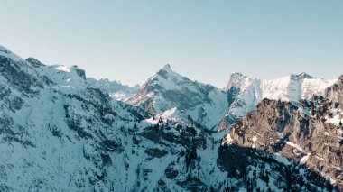 Karla kaplı bir dağ silsilesinin heybetli 4K drone görüntüsü, engebeli zirveler hızla gökyüzüne yükseliyor. Çıtır çıtır beyaz kar manzarayı örtüyor, yukarıdaki derin mavi gökyüzü ile çelişiyor..