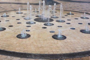 Birden fazla püskürtüsü olan dekoratif bir su çeşmesi içeren dairesel taştan bir plaza. Su çeşitli noktalardan fışkırarak davetkar ve dinamik bir atmosfer yaratır..