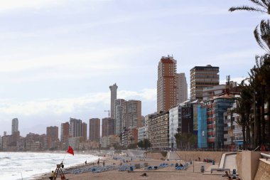 Benidorm, İspanya - 2024.04.27. - Platja del Mal Pas - Sahilde kıyı boyunca uzanan, kumlu sahile dağılmış güneş pansiyonlarıyla dolu canlı bir sahnedir. Dalgalar yavaşça bulutlu bir gökyüzünün altında süzülür.