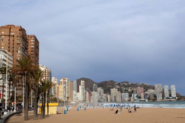 Benidorm, İspanya - 2024.04.27. - Platja de Ponent - Sahilde hareketli bir sahne modern yüksek binaların kıyı boyunca uzandığını gösteriyor. Palmiye ağaçları hafif esintide sallanırken renkli plaj şemsiyeleri bulutlu bir gökyüzünün altında kumlu sahili benekler..
