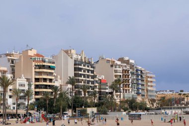 Benidorm, İspanya - 2024.04.27. - Platja del Mal Pas - Palmiyelerin ve çocukların kumsalda oynadığı canlı bir plaj sahnesi. Modern binalar kıyı şeridi boyunca uzanır. Parçalı bulutlu gökyüzüne karşı renkli balkonlar sergilenir..