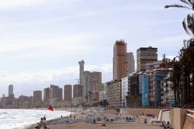 Benidorm, İspanya - 2024.04.27. - Platja del Mal Pas - Sahilde kıyı boyunca uzanan, kumlu sahile dağılmış güneş pansiyonlarıyla dolu canlı bir sahnedir. Dalgalar parçalı bulutlu bir gökyüzünün altında yavaşça yuvarlanarak canlı bir atmosfer yaratıyor..
