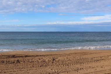 Sakin bir sahil sahnesi, yumuşak dalgaların kumlu kıyıya vurduğu dağınık bulutlu mavi gökyüzü altında sakin bir atmosferi çağrıştırıyor..