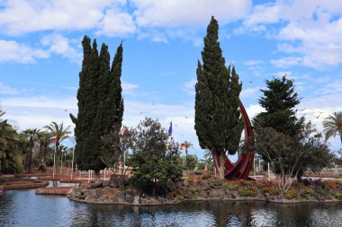 Uzun ağaçlar, canlı çiçekler ve parlak mavi bir gökyüzünün altında yansıtıcı bir gölet olan sakin bir parkın manzarası, dingin bir doğal vaha yaratıyor..