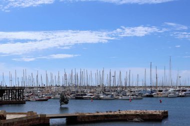 Durgun sulara kenetlenmiş sayısız yatın bulunduğu sakin bir liman sahnesi bulutlu berrak bir mavi gökyüzüne karşı kurulmuş, barışçıl ve davetkar bir deniz atmosferi yaratıyor..