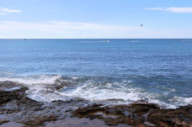 Uçsuz bucaksız mavi denizin çarpıcı manzarası ve dalgaları ile kayalık bir sahil kıyısı. Uzakta birkaç tekne, kıyı yaşamının ve maceranın güzelliğini kapsıyor..