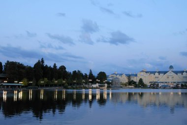 Disneyland Paris, Fransa -2024.06.05.Newport Körfezi Kulübü A sakin gölü çevresindeki binaların yumuşak ışıklarını yansıtır. Sakin sular ve uzak ağaçlar huzurlu bir atmosfer yaratarak izleyicileri doğanın dingin güzelliğinin tadını çıkarmaya çağırıyor..
