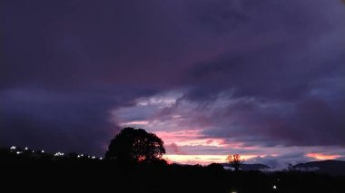 Gökyüzünün turuncu ve pembe renkli, koyu ve dramatik bulutlarla çelişen canlı tonlarıyla boyandığı muhteşem bir günbatımı. Ağaç siluetleri göze çarpıyor, uzak ışıklar parıldarken, gizem ve sükunet katıyor..