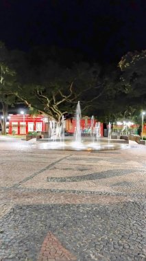 Gece sessiz bir meydanın ortasında, ağaçlarla çevrili ve taşlarla döşenmiş güzel aydınlatılmış bir çeşme. Atmosfer huzur ve sükunet hissi uyandırıyor. Gece yürüyüşü için mükemmel..
