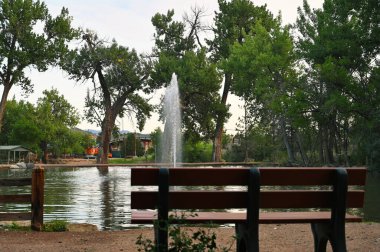 Park göletinin ortasındaki bir çeşme arka planda ağaçlar olan bir bankın arkasından görülüyor..