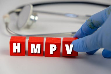 Üzerinde HMPV yazılı bir levha tutan eldiven giymiş bir sağlık görevlisinin resmi. Arka planda steteskop ve tıbbi maske var. İnsan metafnömotorirüsünün (HMPV) belirtileri ciddiyette değişebilir.
