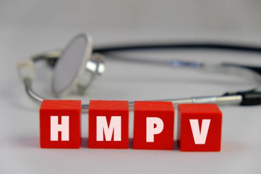 Üzerinde HMPV yazılı bir levha tutan eldiven giymiş bir sağlık görevlisinin resmi. Arka planda steteskop ve tıbbi maske var. İnsan metafnömotorirüsünün (HMPV) belirtileri ciddiyette değişebilir.