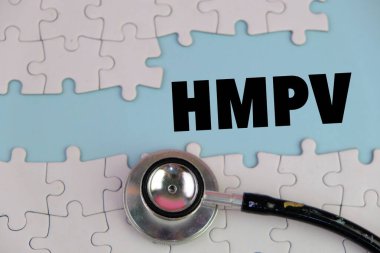 Üzerinde HMPV yazılı bir levha tutan eldiven giymiş bir sağlık görevlisinin resmi. Arka planda steteskop ve tıbbi maske var. İnsan metafnömotorirüsünün (HMPV) belirtileri ciddiyette değişebilir.