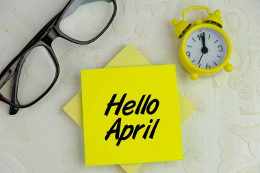 Merhaba April. Beyaz arkaplandaki sarı not defteri kağıdına yazı. Alarm saatinin yanında
